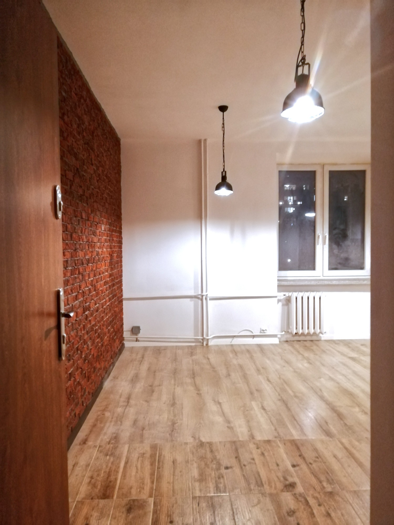 Pusty pokój po remoncie z drewnianą podłogą, ścianą z czerwonej cegły i dwoma lampami wiszącymi, widok przez otwarte drzwi.