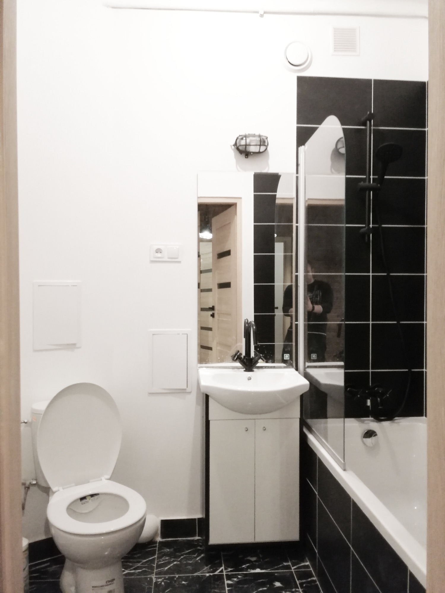 Nowoczesna łazienka z czarno-białymi płytkami, wiszącą szafką z umywalką, toaletą i kabiną prysznicową z czarną armaturą; widoczne detale wykończenia, takie jak oświetlenie i wentylacja.