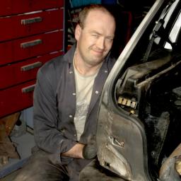 AUTO-SERVICE Jerzy Łęczycki - Uśmiechnięty mechanik w warsztacie, siedzący obok otwartego bagażnika samochodu, w tle czerwona szafka narzędziowa.