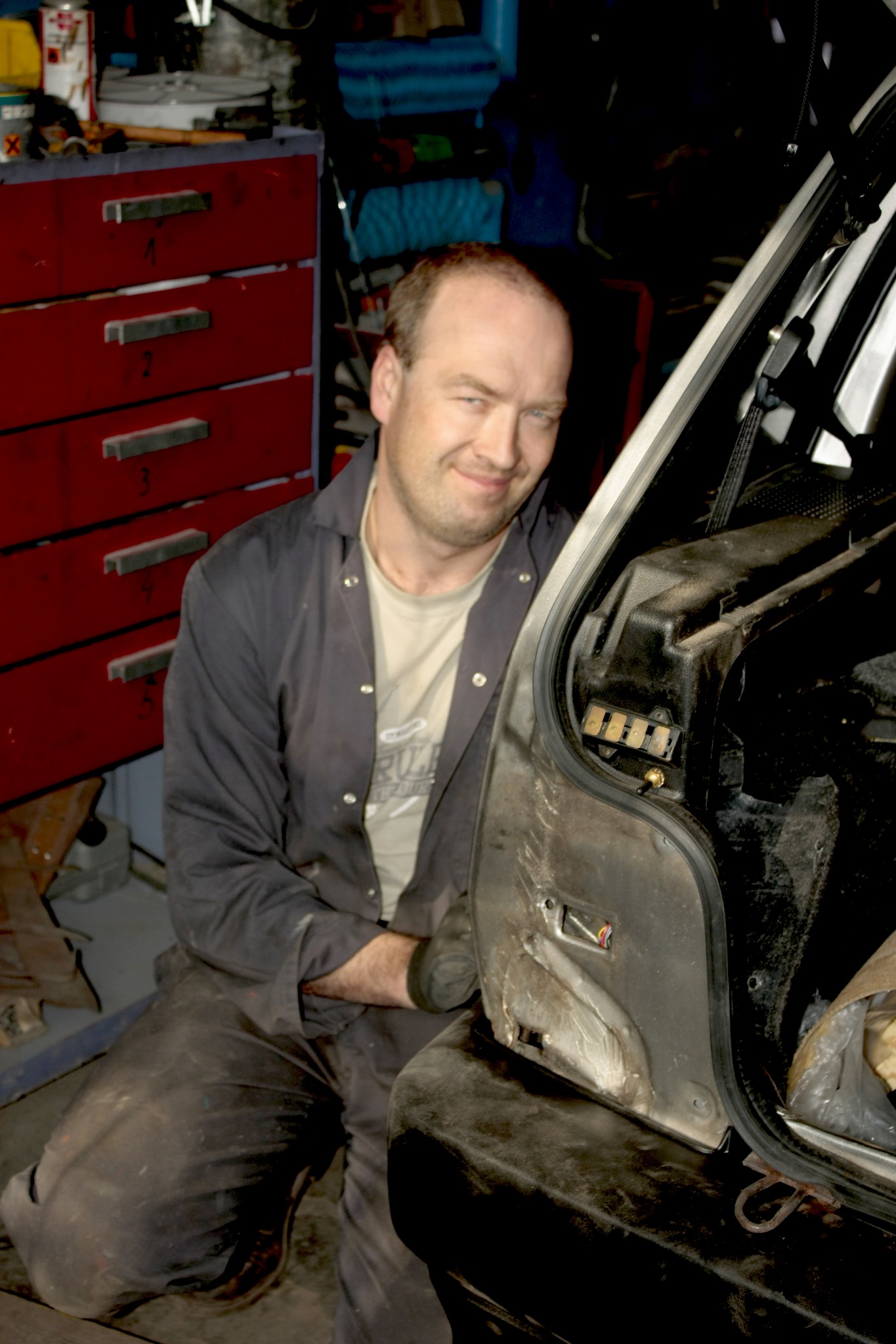 Uśmiechnięty mechanik w warsztacie, siedzący obok otwartego bagażnika samochodu, w tle czerwona szafka narzędziowa.