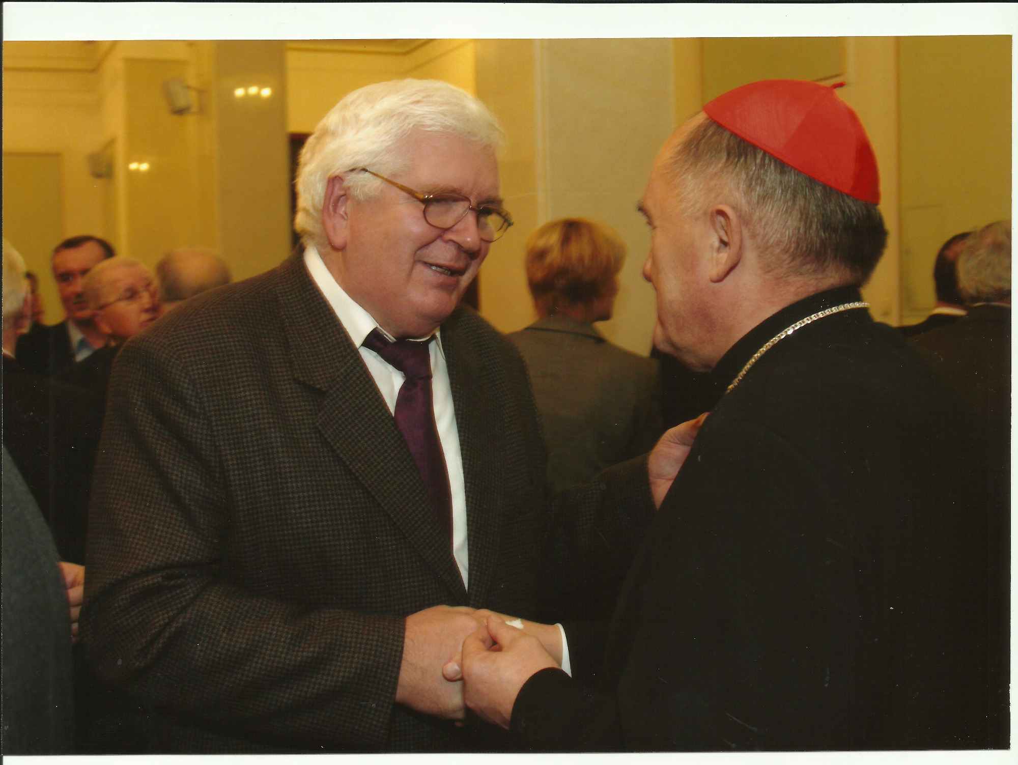 Uśmiechnięty mężczyzna w brązowym tweedowym garniturze i okularach ściska dłoń mężczyzny w czarnej sutannie i czerwonej piusce w jasnym wnętrzu.