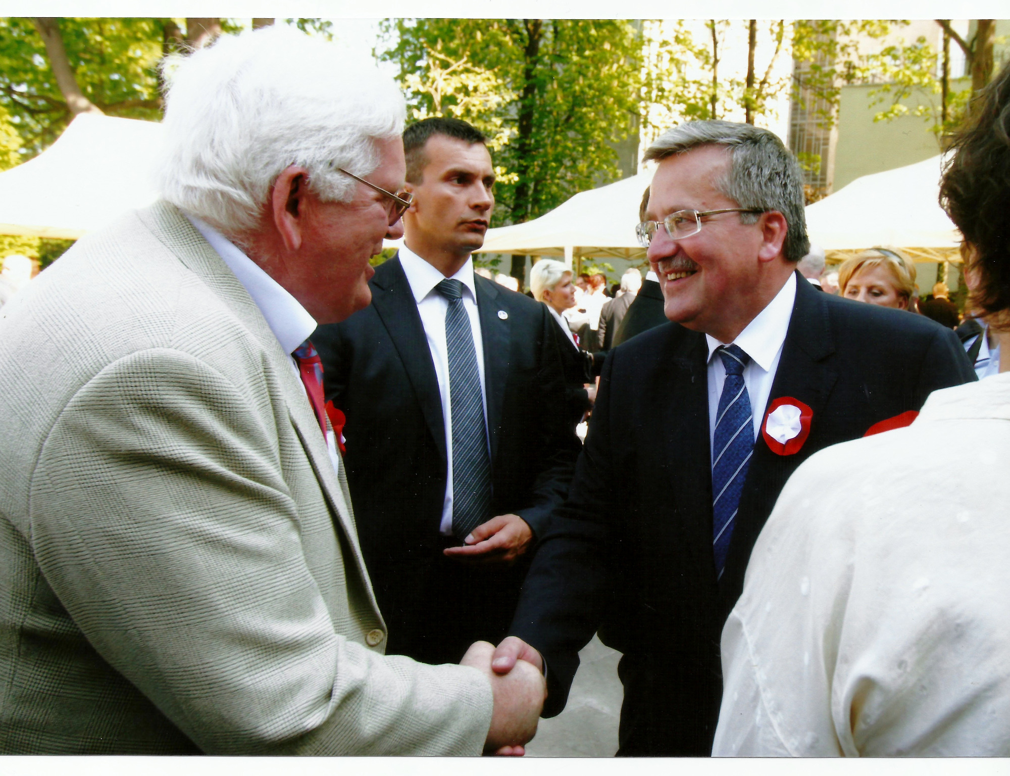 Dwóch uśmiechniętych mężczyzn w garniturach w trakcie uścisku dłoni na tle zieleni i białych namiotów.