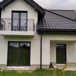 Nowoczesny dom jednorodzinny z czarnymi oknami i balkonem z ozdobną, kutą balustradą, widok z zewnątrz na elewację, w oknach odbija się zieleń ogrodu.