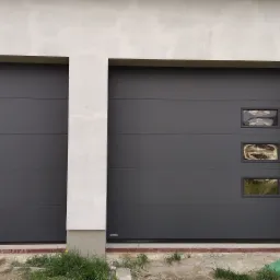 Dwa garaże z szarymi segmentowymi bramami garażowymi, jedna z trzema małymi okienkami, ściany garażu w surowym tynku.