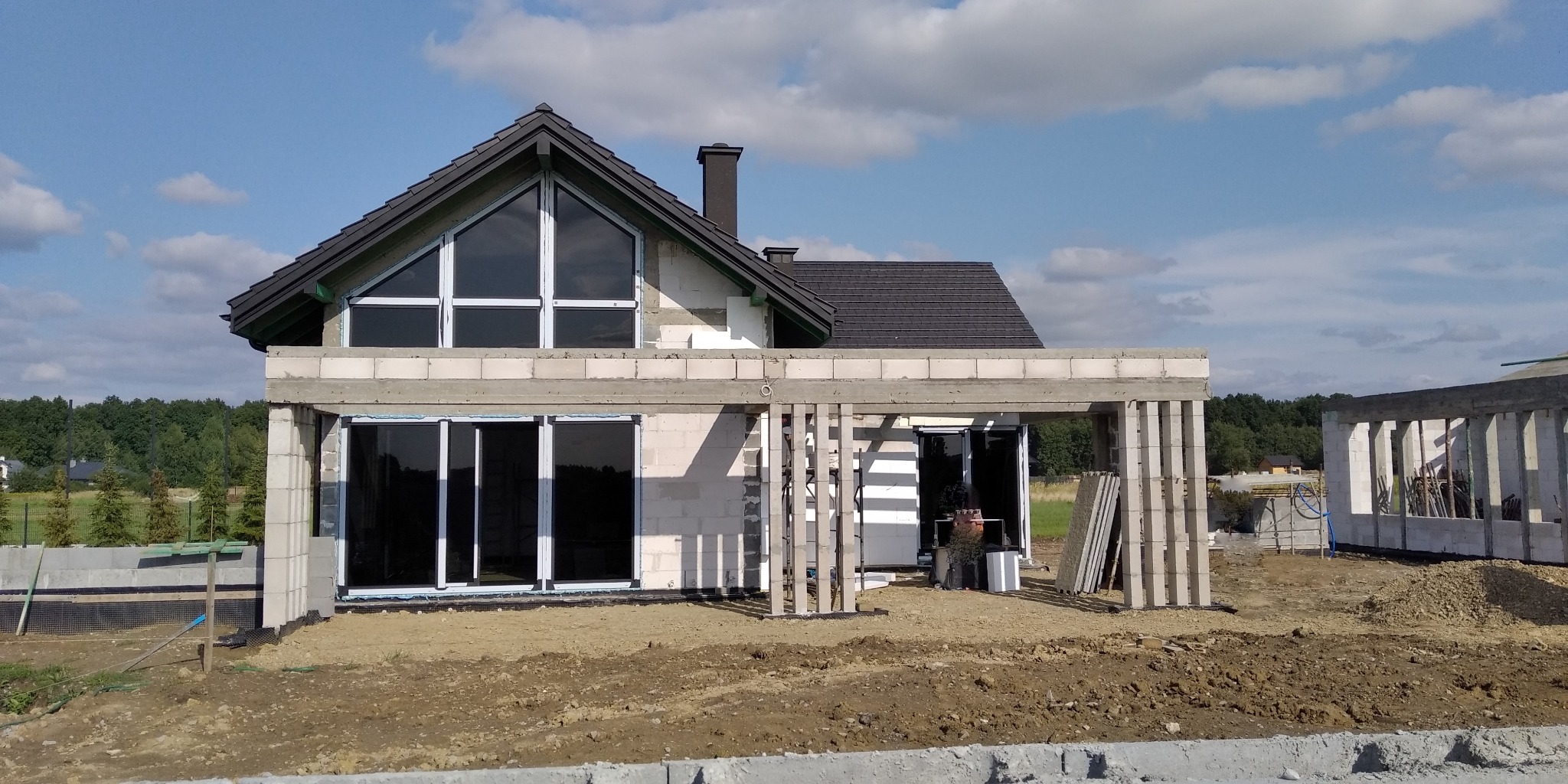 Surowy stan budowy domu jednorodzinnego z zamontowanymi oknami PCV i przesuwnymi drzwiami tarasowymi, widoczne bloczki betonowe i niezabezpieczony teren wokół budynku.