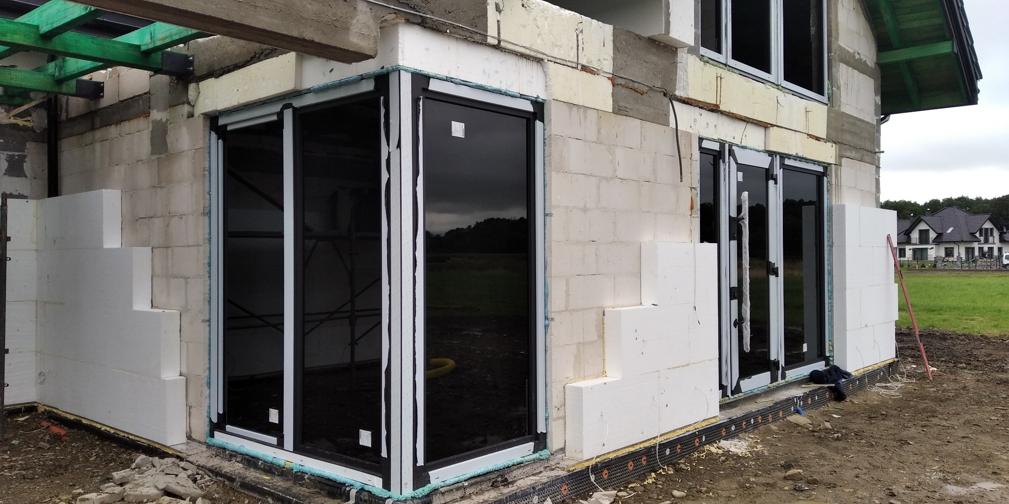 Montaż dużych przesuwnych drzwi tarasowych z czarnymi ramami w surowym stanie budynku, widoczne ocieplenie styropianem i niezabudowany teren.