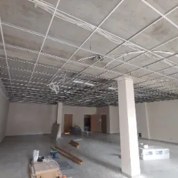 Metalowa konstrukcja sufitu podwieszanego z widocznymi przewodami elektrycznymi w przestronnym, jasnym pomieszczeniu biurowym w trakcie remontu, z materiałami budowlanymi na podłodze.