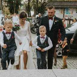 Joanna Adamczak Fotografia - Para młoda z dwójką chłopców w eleganckich strojach wychodząca z kościoła po ceremonii ślubnej, w tle goście i zaparkowane samochody.