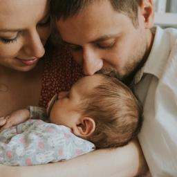 Joanna Adamczak Fotografia - Mama i tata tulą śpiące niemowlę, tata całuje dziecko w główkę, spokojna atmosfera rodzinna, pastelowe kolory.