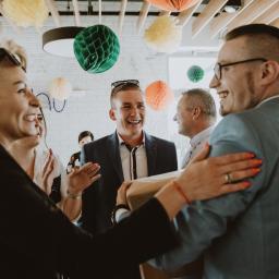 Joanna Adamczak Fotografia - Uśmiechnięci goście w eleganckich strojach witają parę trzymającą prezent, dekoracje z papierowych kul wiszą pod sufitem.