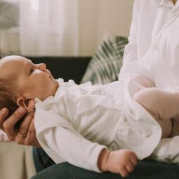 Joanna Adamczak Fotografia - Noworodek w białej sukience i różowych skarpetkach leży na rękach osoby w białej koszuli, widoczny złoty pierścionek na palcu.