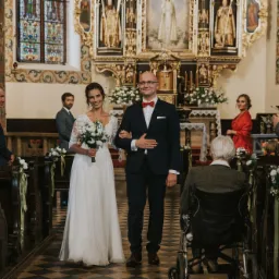 Para młoda idzie alejką w kościele, panna młoda w białej sukni z bukietem, pan młody w granatowym garniturze z czerwoną muchą, w tle ołtarz i goście.