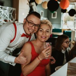 Joanna Adamczak Fotografia - Uśmiechnięta para: mężczyzna w okularach, muszce i szelkach obejmuje kobietę w czerwonej sukience, trzymającą kieliszek z napojem, w tle dzieci przy stole i dekoracje z papierowych kul.