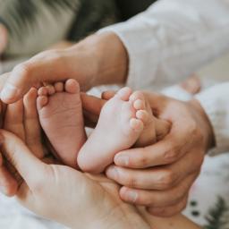 Joanna Adamczak Fotografia - Dłonie rodziców delikatnie obejmują stópki noworodka na tle kwiecistej pościeli, ukazując intymność i więź rodzinną.