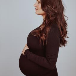 Joanna Adamczak Fotografia - Kobieta w obcisłej sukience w kolorze burgundu, z rękami na dużym brzuchu ciążowym, profil z zamkniętymi oczami i długimi, falowanymi, brązowymi włosami na jasnoszarym tle.