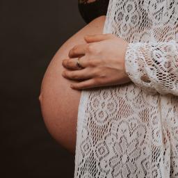 Joanna Adamczak Fotografia - Zaokrąglony brzuch kobiety w ciąży, dłoń z złotą obrączką spoczywająca na brzuchu, okryty ażurową białą narzutką na ciemnym tle.