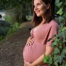 Joanna Adamczak Fotografia - Uśmiechnięta kobieta w różowej sukience, trzymająca ręce na brzuchu, pozuje na tle zieleni parku z widocznym mostkiem nad wodą.