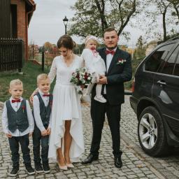 Joanna Adamczak Fotografia - Para młoda z dwoma synami i córeczką w białej sukience, pozują na brukowanej drodze obok czarnego SUV-a, bukiet ślubny w dłoniach panny młodej.