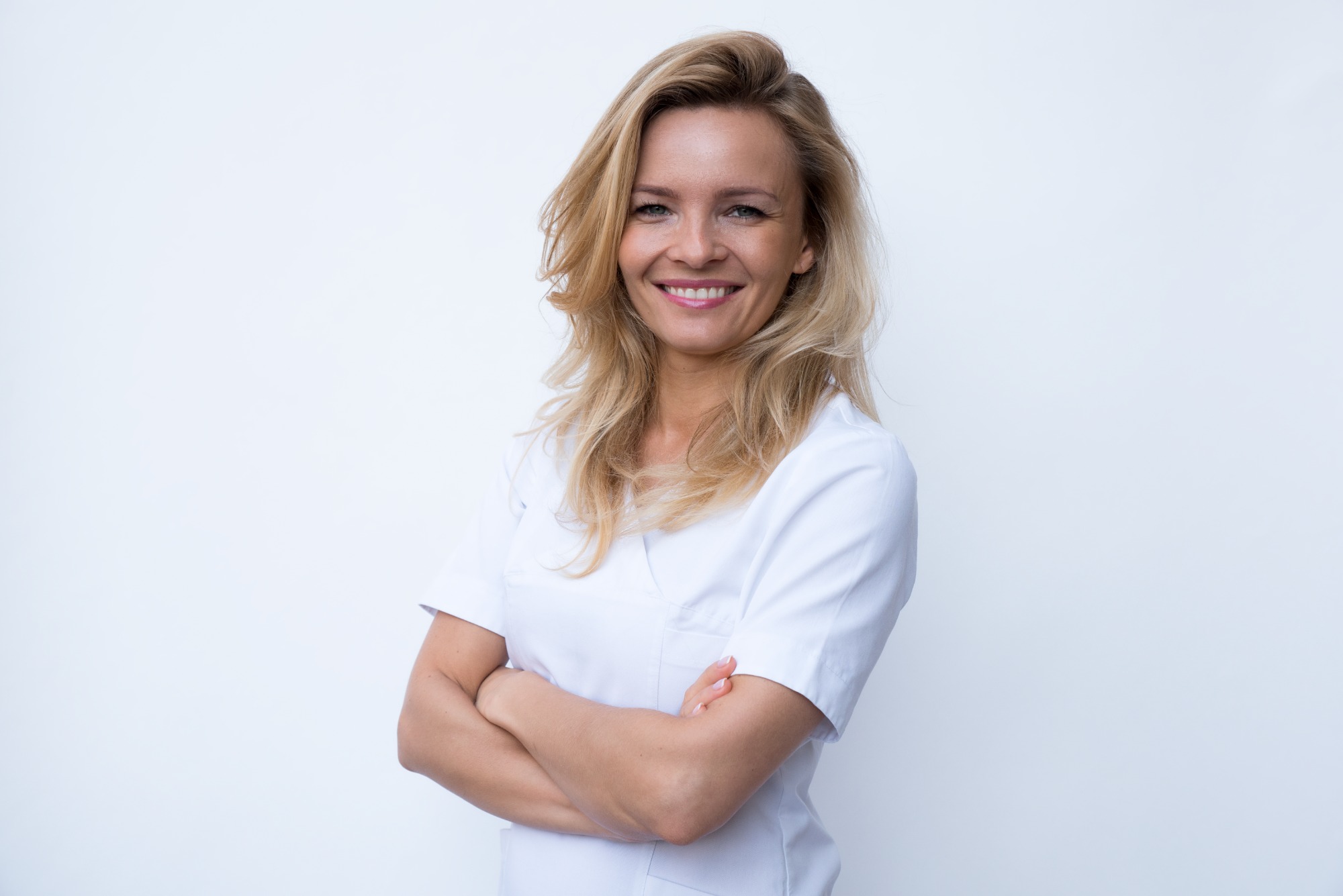 Portret uśmiechniętej blondynki w białym uniformie na jasnym tle, ręce skrzyżowane na piersiach.