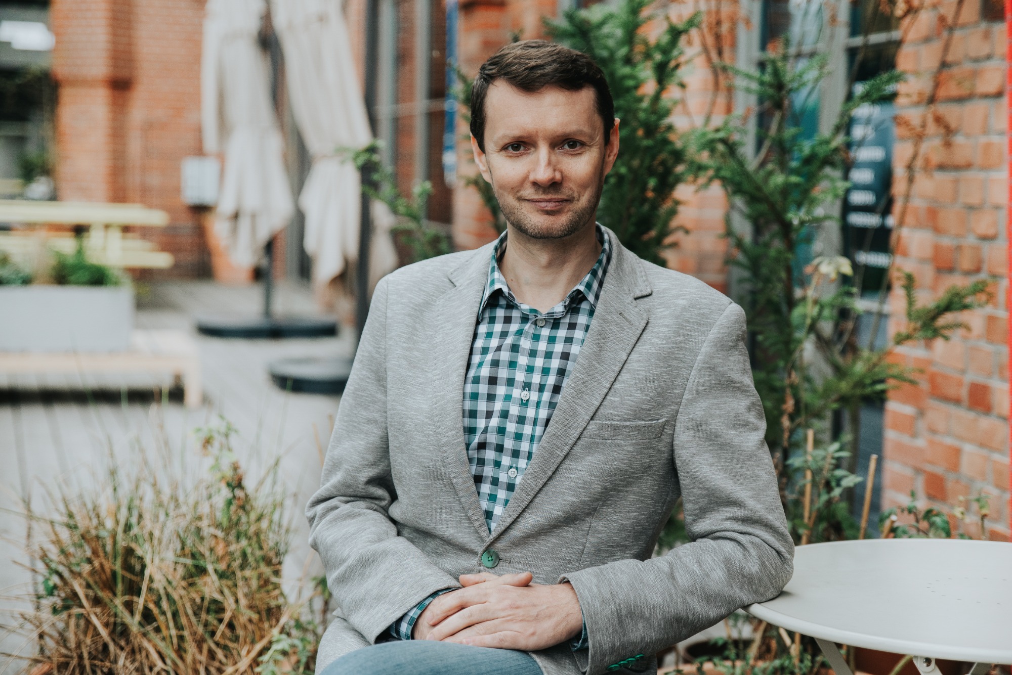 Portret uśmiechniętego mężczyzny w szarej marynarce i kraciastej koszuli, siedzącego przy białym stoliku na tle ceglanej ściany i zieleni.