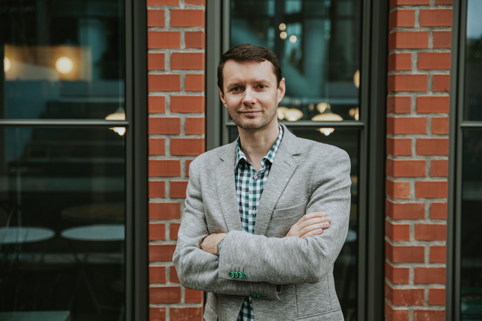 Portret mężczyzny w szarej marynarce i kraciastej koszuli, stojącego na tle ściany z czerwonej cegły i okna z ciemną ramą.