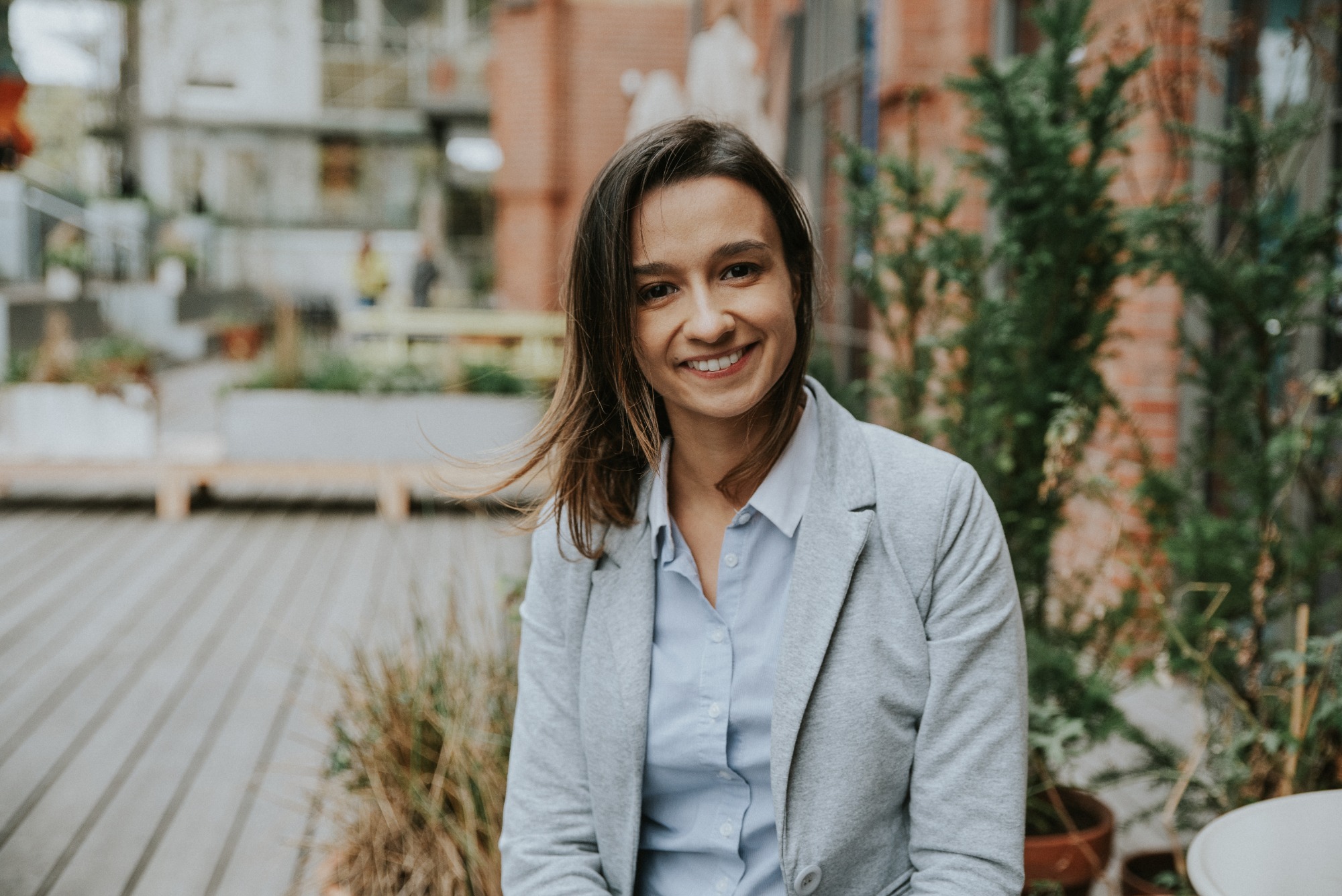 Uśmiechnięta kobieta w szarej marynarce i błękitnej koszuli, pozująca na tle miejskiej przestrzeni z zielenią i budynkami z czerwonej cegły.