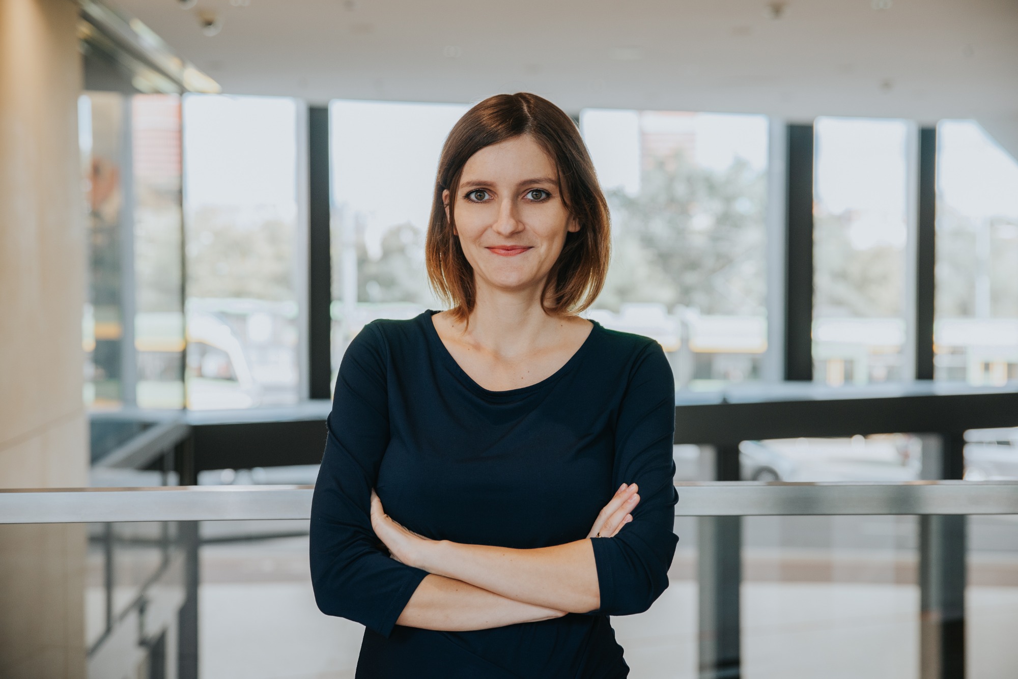 Kobieta z brązowymi włosami w ciemnej bluzce, z założonymi rękami na tle przeszklonej przestrzeni biurowej.