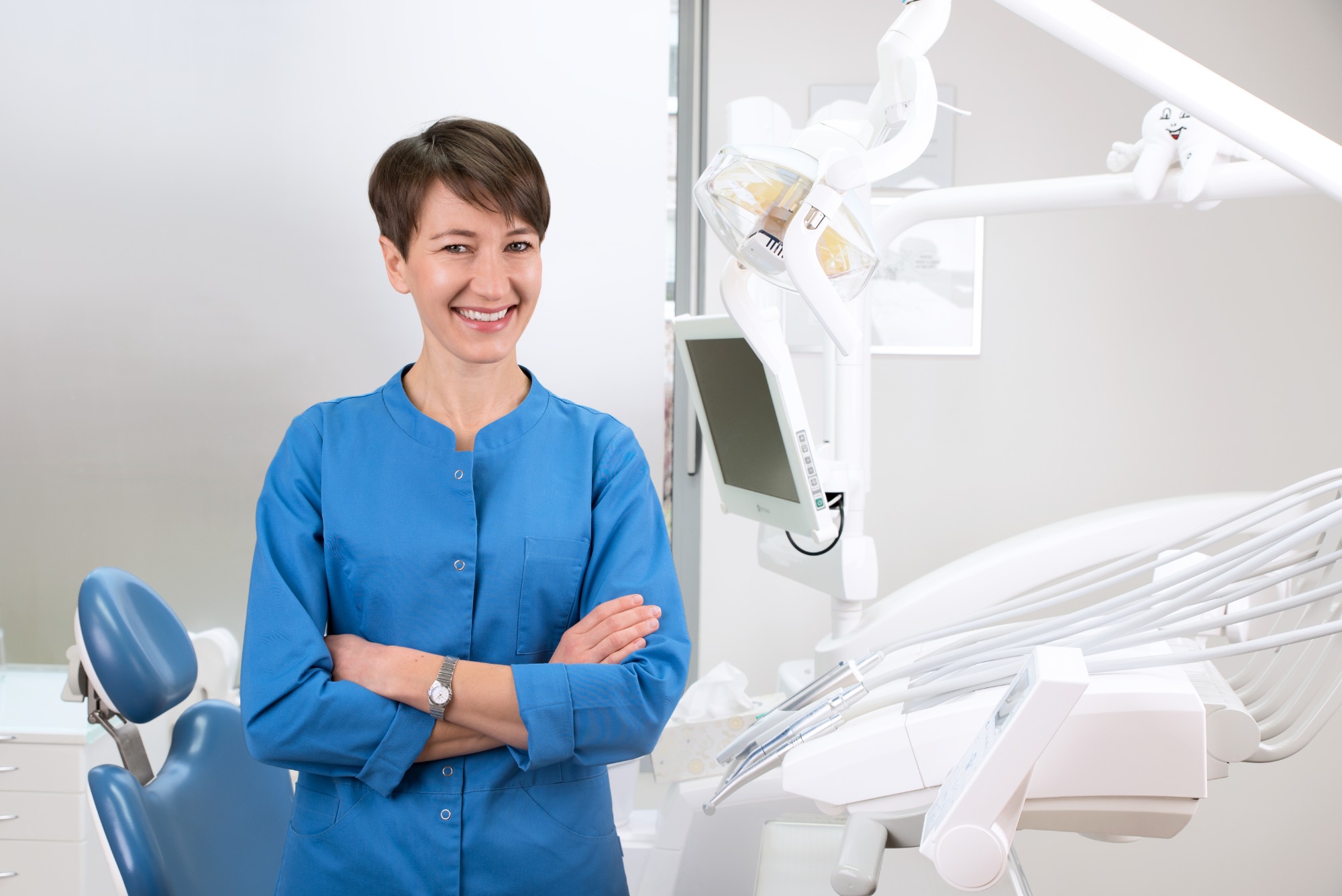 Uśmiechnięta dentystka w niebieskim uniformie, z zegarkiem na ręku, stoi na tle nowoczesnego gabinetu stomatologicznego z fotelem i specjalistycznym sprzętem.