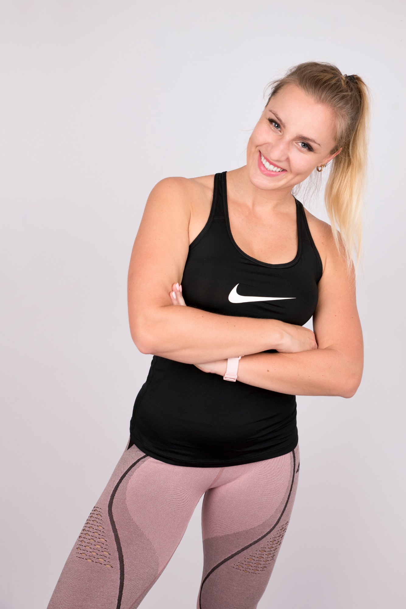 Uśmiechnięta blondynka w czarnym topie sportowym z logo Nike i różowych legginsach, stojąca na jasnym tle z założonymi rękami.