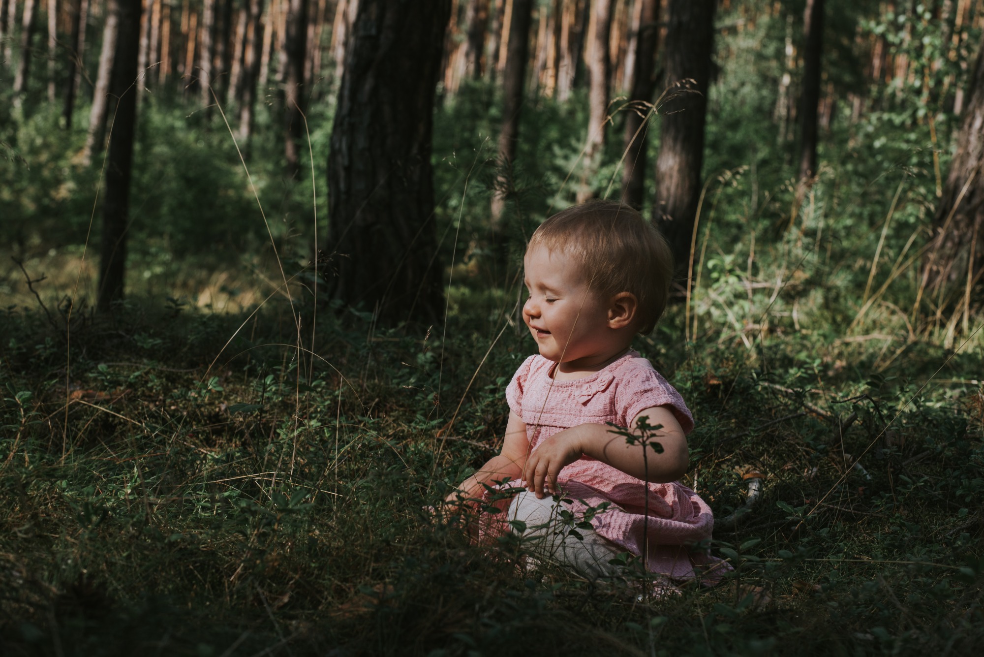 Uśmiechnięta dziewczynka w różowej sukience siedzi w trawie na tle leśnych drzew.