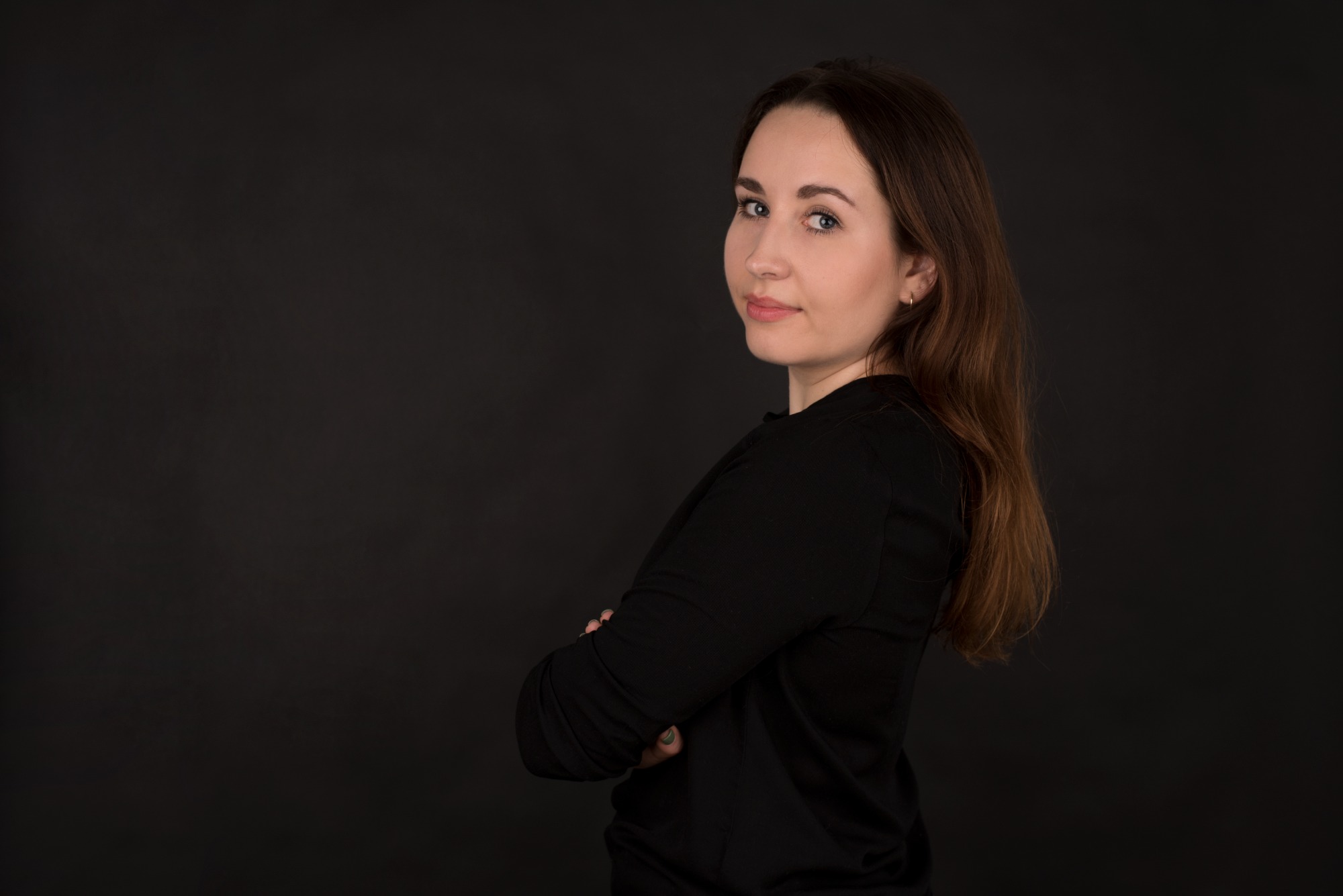Portret kobiety z brązowymi włosami, ubrana w czarną bluzkę, pozująca na ciemnym tle z rękami skrzyżowanymi na piersiach i patrząca przez ramię.
