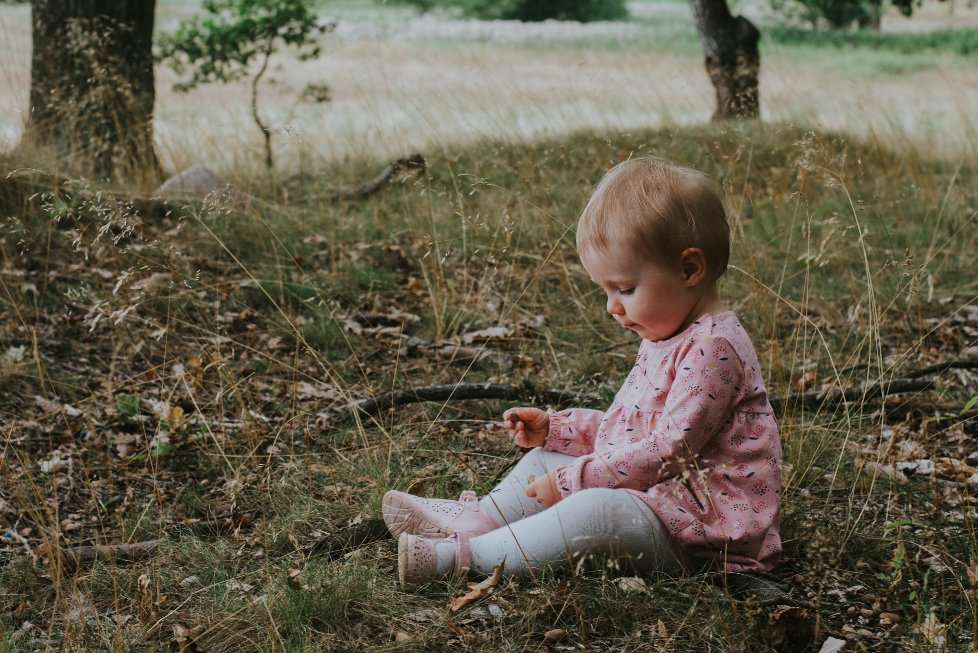 Dziewczynka w różowej sukience siedzi w trawie w Poznaniu, skupiona na małym kamyku w dłoni, delikatne światło podkreśla naturalność sceny.