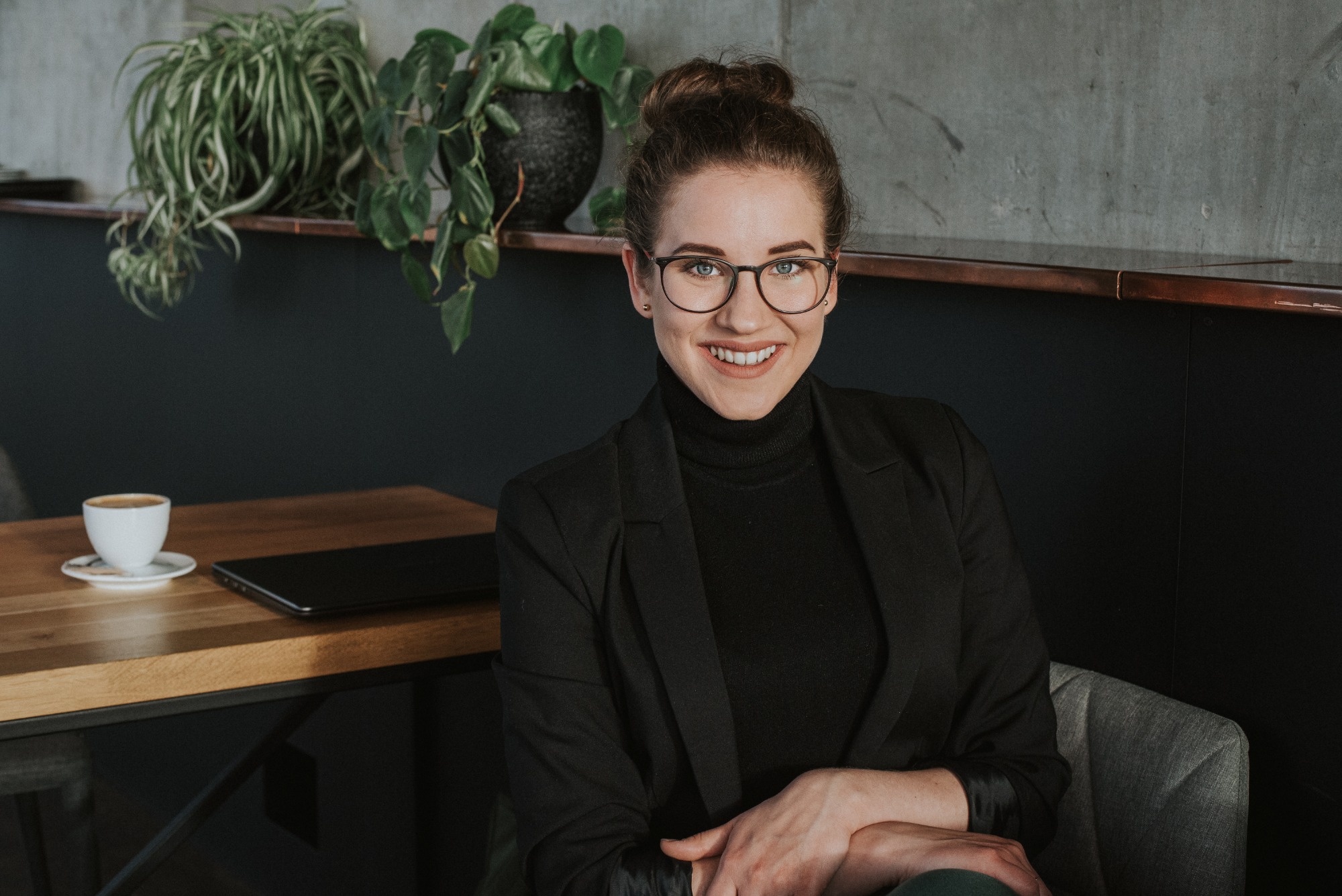 Portret uśmiechniętej kobiety w okularach i czarnym żakiecie, siedzącej w kawiarni z filiżanką kawy i laptopem na stole, rośliny doniczkowe w tle.