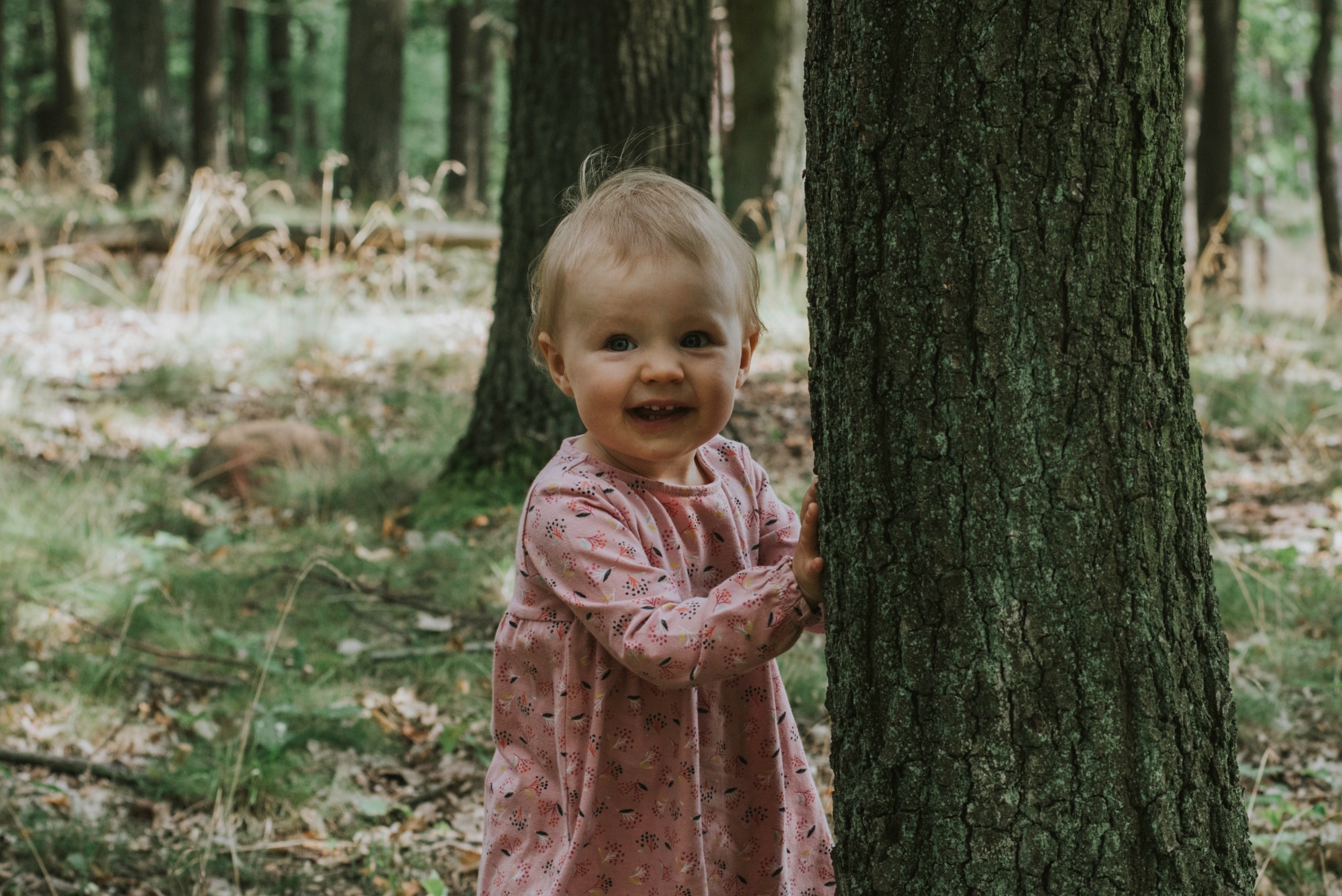 Uśmiechnięta dziewczynka w różowej sukience z kwiatowym wzorem przytula się do grubego drzewa w lesie, z widoczną fakturą kory i zielonym mchem.