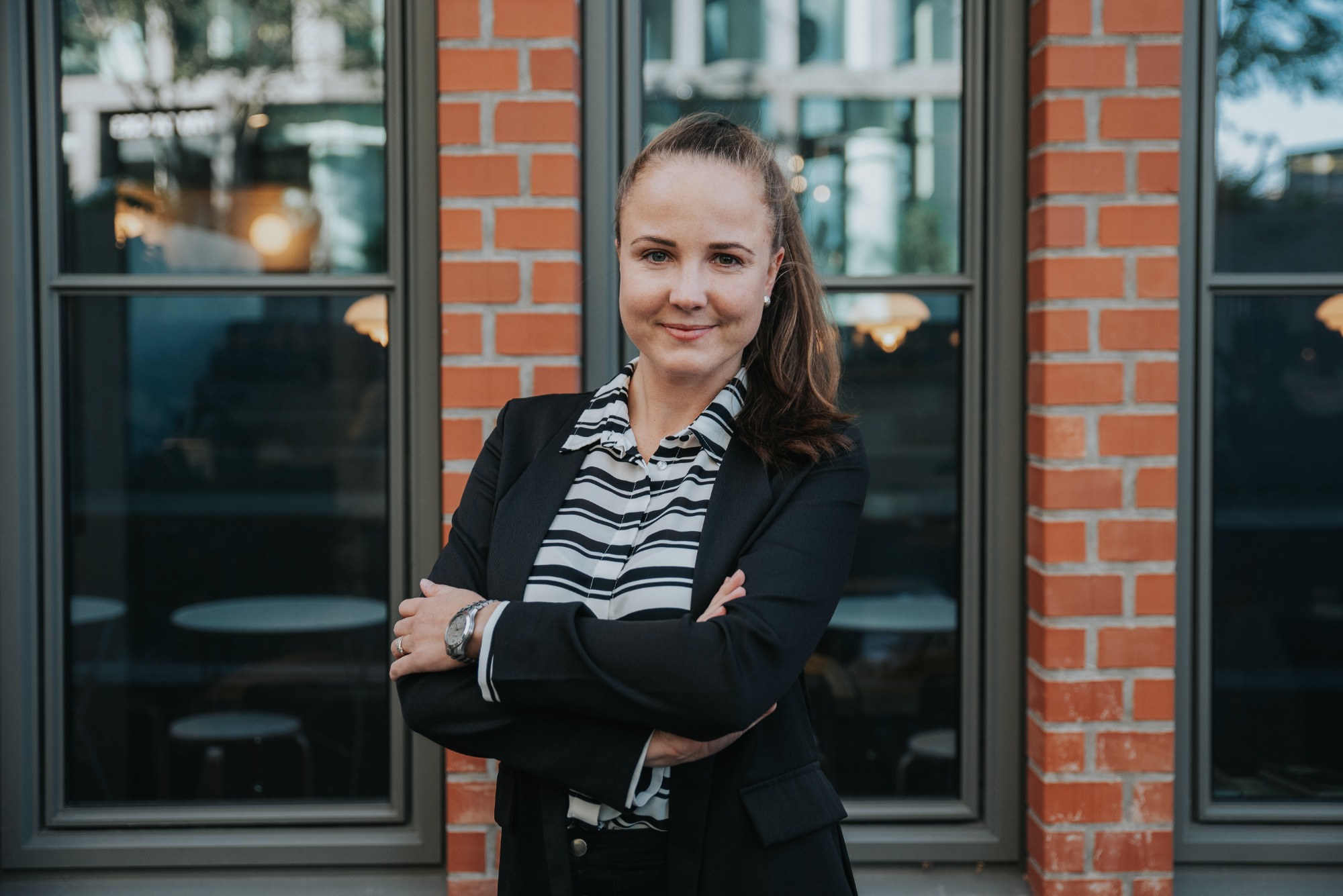 Portret kobiety w czarnym żakiecie i bluzce w paski, z założonymi rękami, na tle ceglanej ściany z oknami.