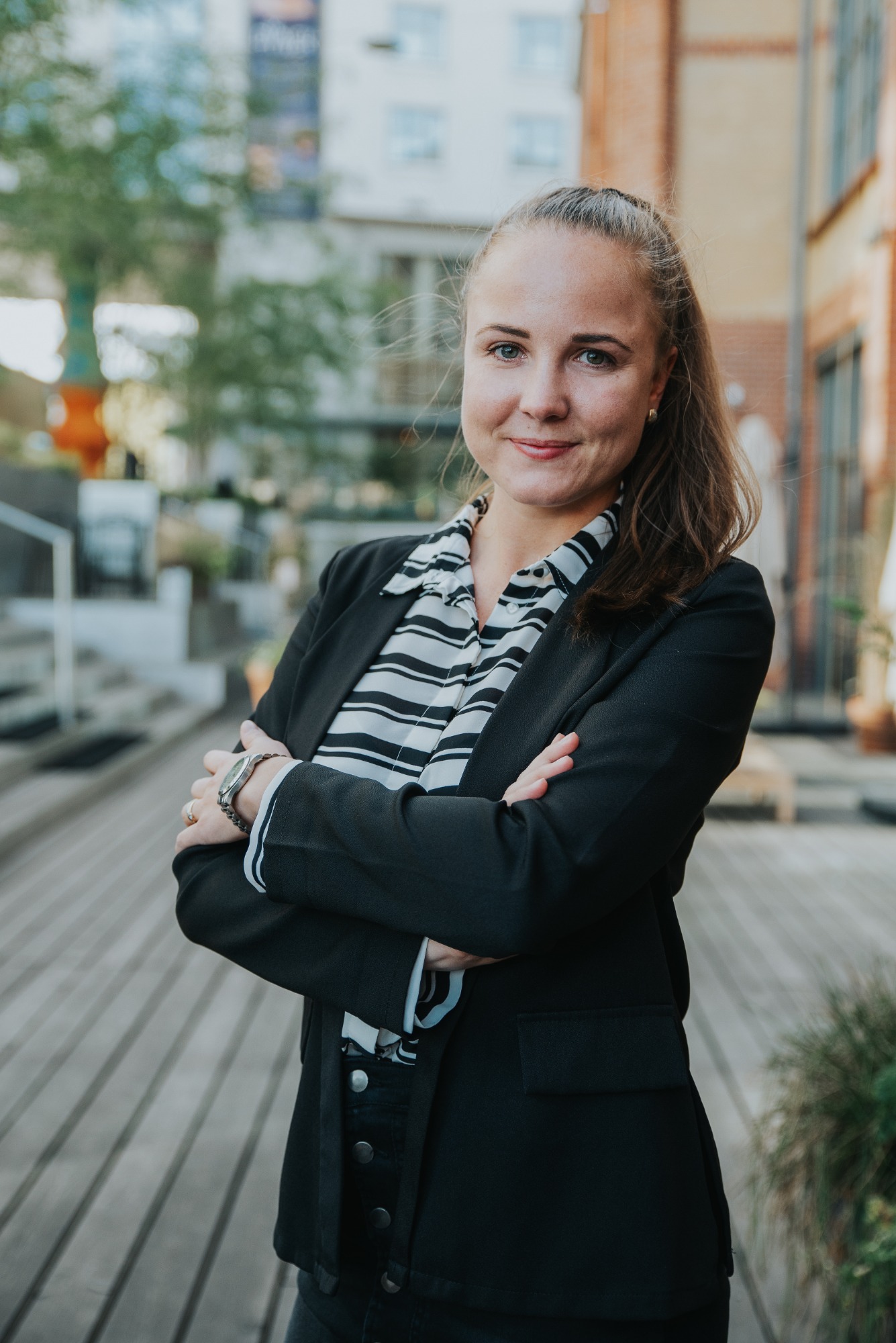 Uśmiechnięta kobieta w czarnej marynarce i koszuli w paski, z założonymi rękami na tle miejskiej przestrzeni z drewnianym podestem.