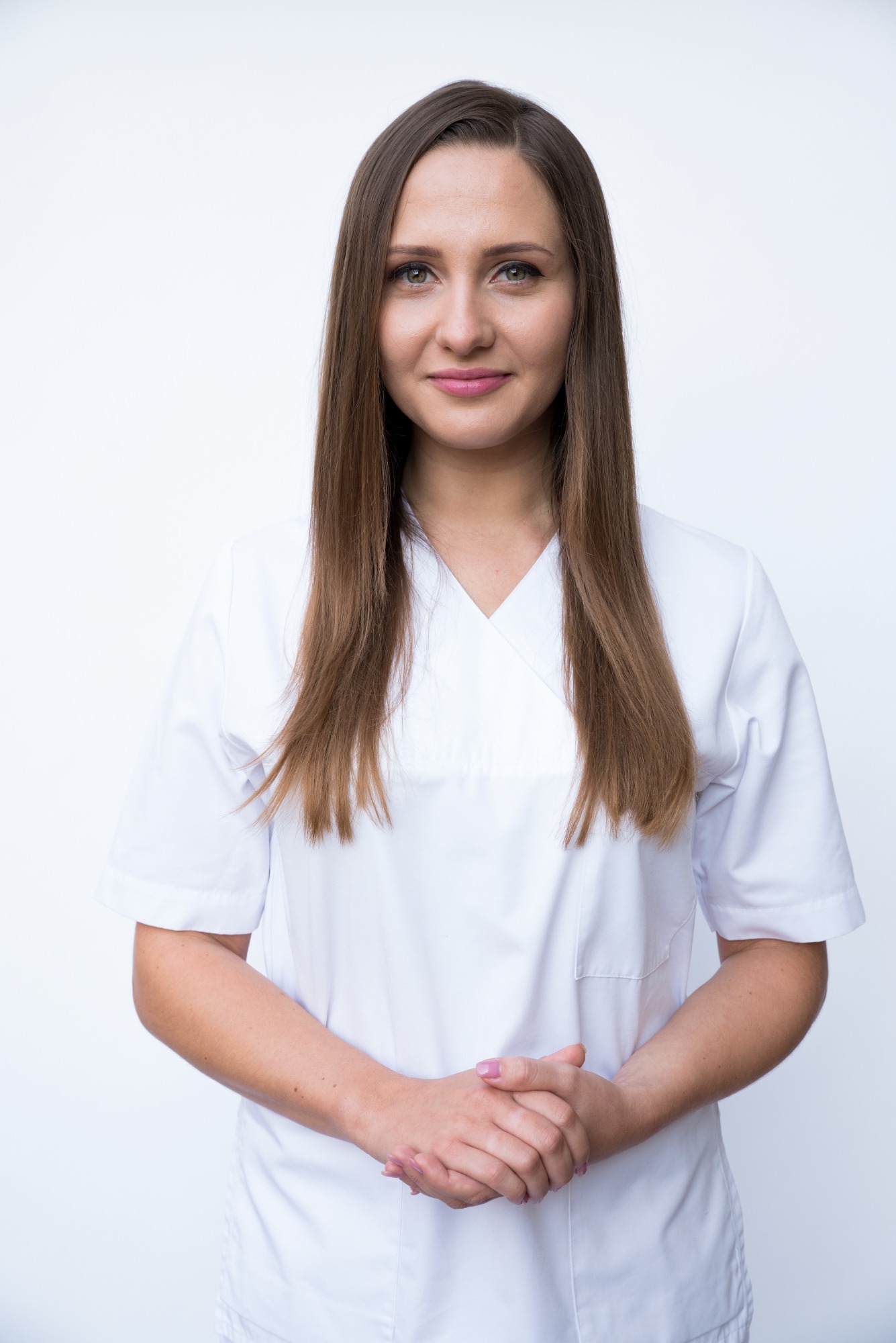 Portret kobiety w białym uniformie medycznym na jasnym tle, z rękami splecionymi przed sobą.