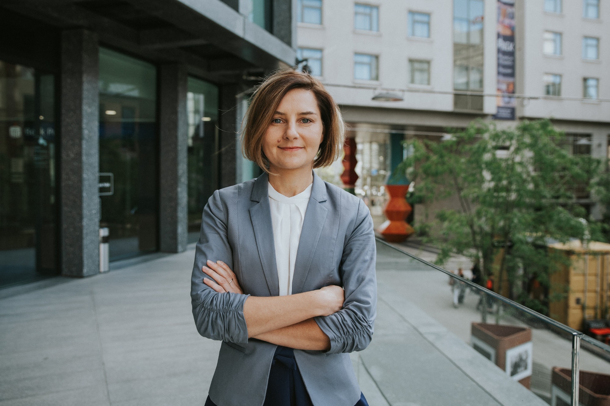 Portret kobiety w szarym żakiecie, stojącej na tle nowoczesnego budynku z przeszklonymi elementami i drzewami, skrzyżowane ramiona, uśmiech.