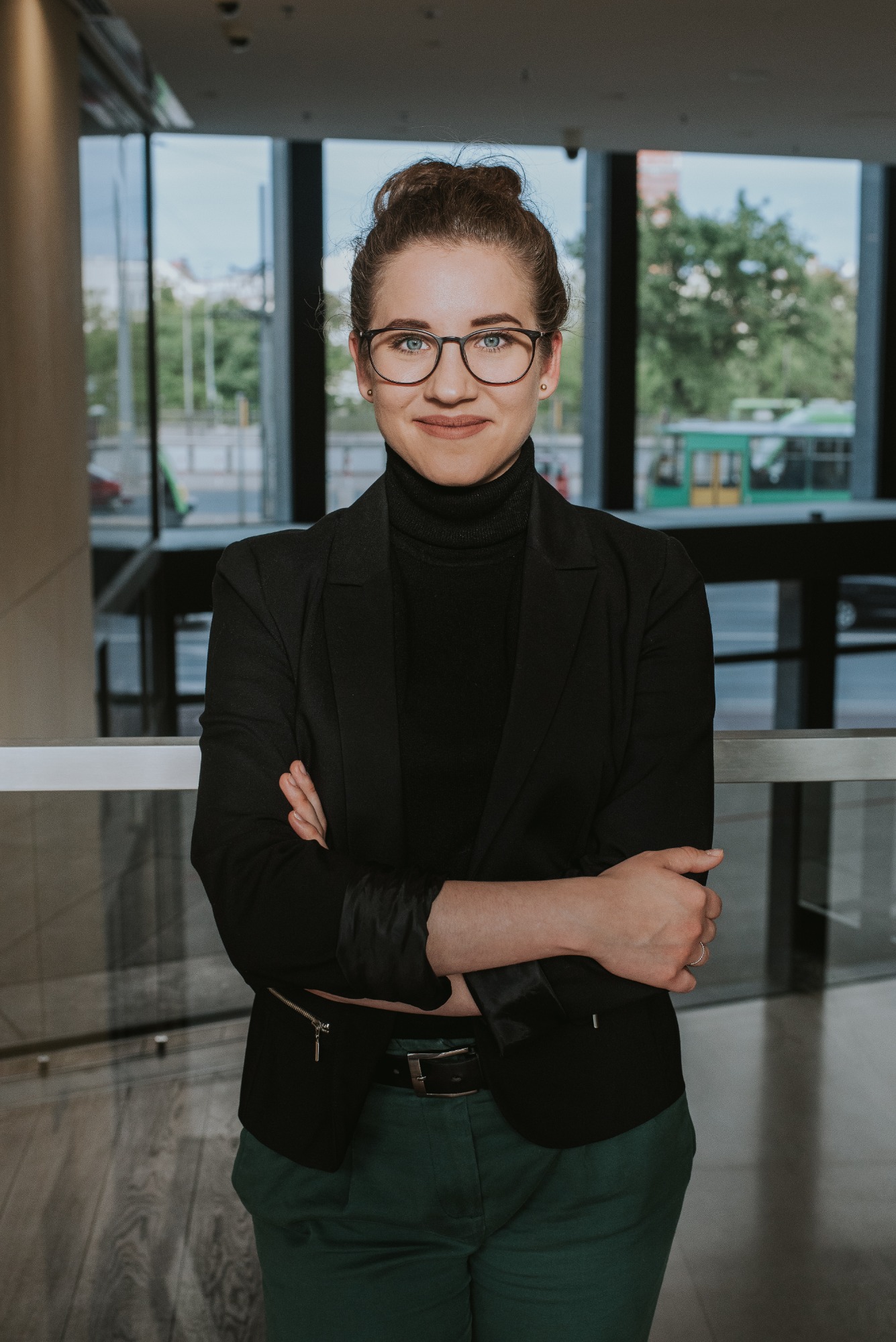 Portret kobiety w okularach, czarnym golfie i marynarce, z założonymi rękami na tle przeszklonego budynku z widokiem na ulicę.