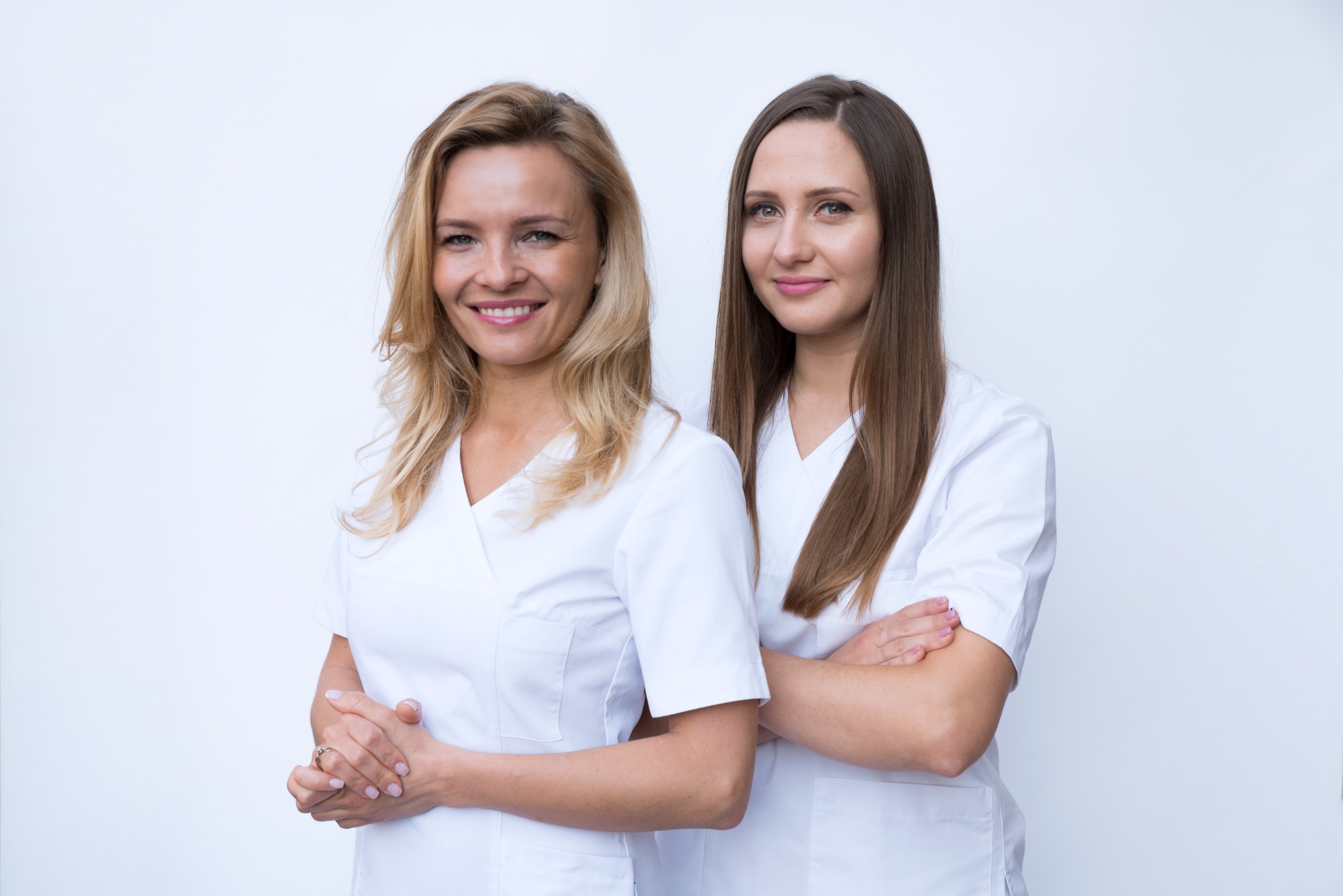 Dwie uśmiechnięte kobiety w białych uniformach stoją na jasnym tle, jedna z blond włosami i pierścionkiem, druga z ciemnymi włosami i skrzyżowanymi ramionami.