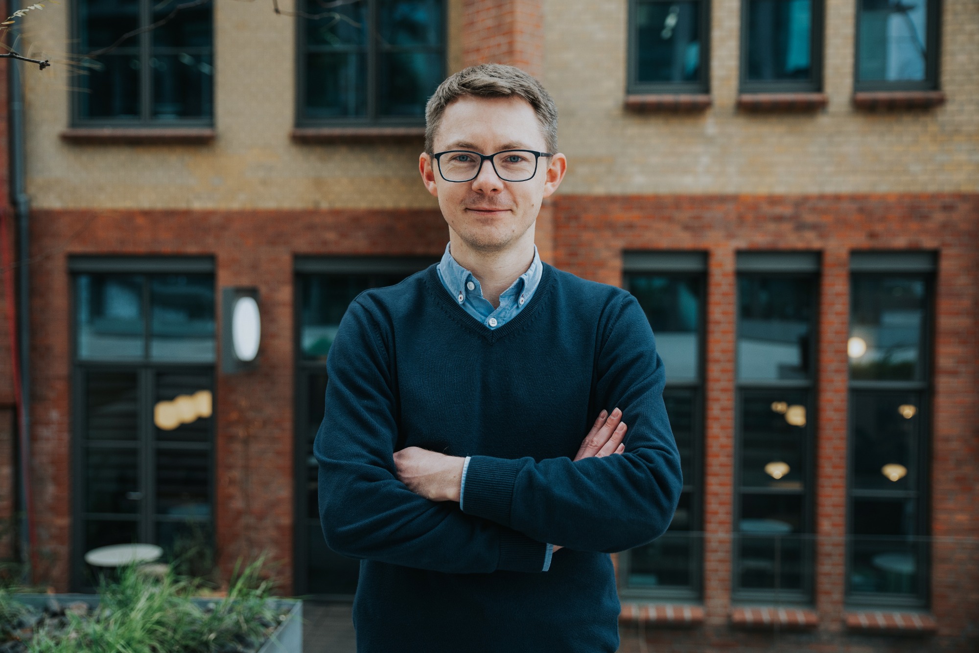 Portret mężczyzny w okularach, ubranego w niebieski sweter i koszulę, stojącego na tle ceglanego budynku z dużymi oknami.