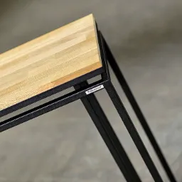 Fragment stołu z blatem z jasnego drewna i czarną metalową ramą z naklejoną etykietą 'StalConcept', na tle szarej, betonowej podłogi