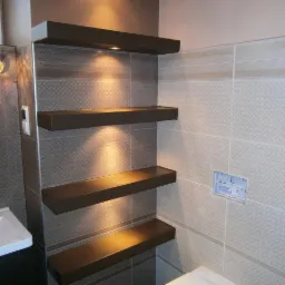 Wykończona łazienka z półkami podświetlanymi punktowo, płytki w odcieniach szarości i beżu. Widoczna umywalka i fragment toalety.