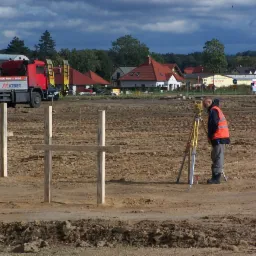 Pracownik w pomarańczowej kamizelce używa niwelatora na placu budowy z drewnianymi słupkami, w tle czerwona ciężarówka MATBET i budynki mieszkalne pod pochmurnym niebem.