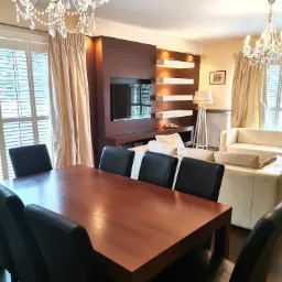 Jasne wnętrze salonu z drewnianym stołem, czarnymi krzesłami, kremową sofą i kryształowymi żyrandolami, oświetlone światłem dziennym wpadającym przez żaluzje.