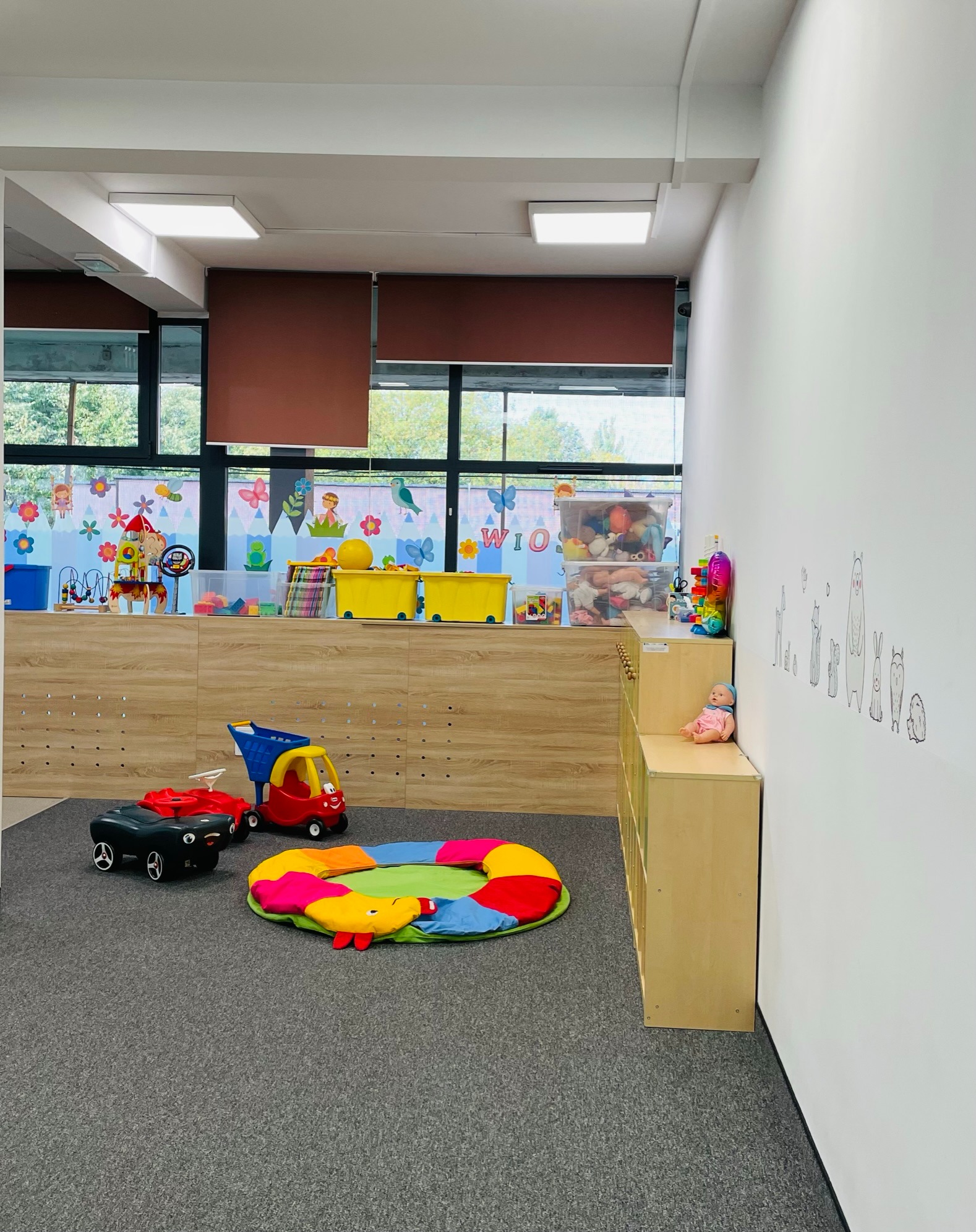 Jasny pokój zabaw z zabawkami: kolorowa mata, plastikowe pojazdy i półki z zabawkami, okno z dekoracjami i rysunki zwierząt na ścianie.