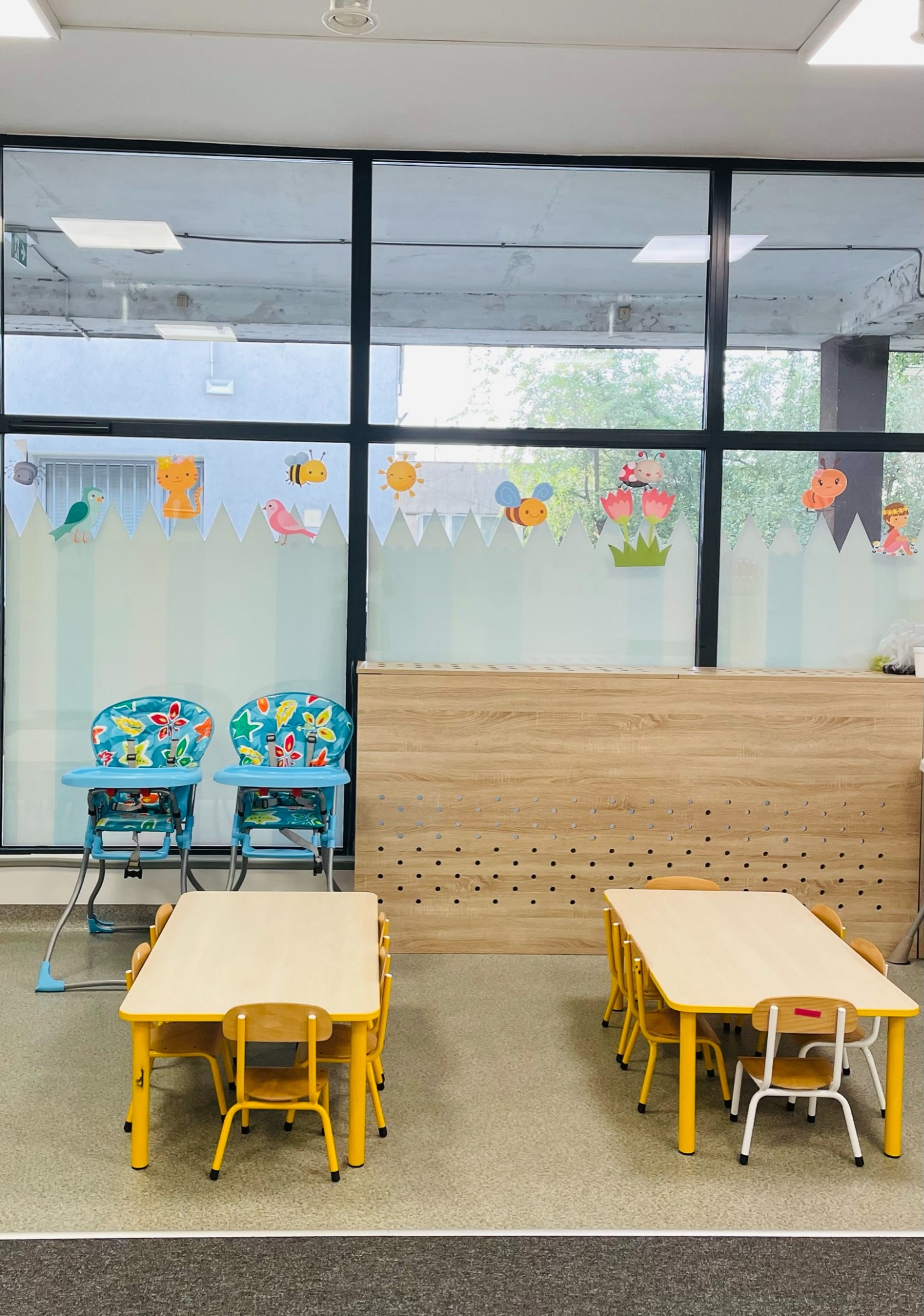 Sala z dwoma stolikami z żółtymi nogami i krzesłami oraz dwoma krzesełkami do karmienia z niebieskimi ramami i kolorowymi siedziskami, okno ozdobione naklejkami z motywami zwierząt i roślin.