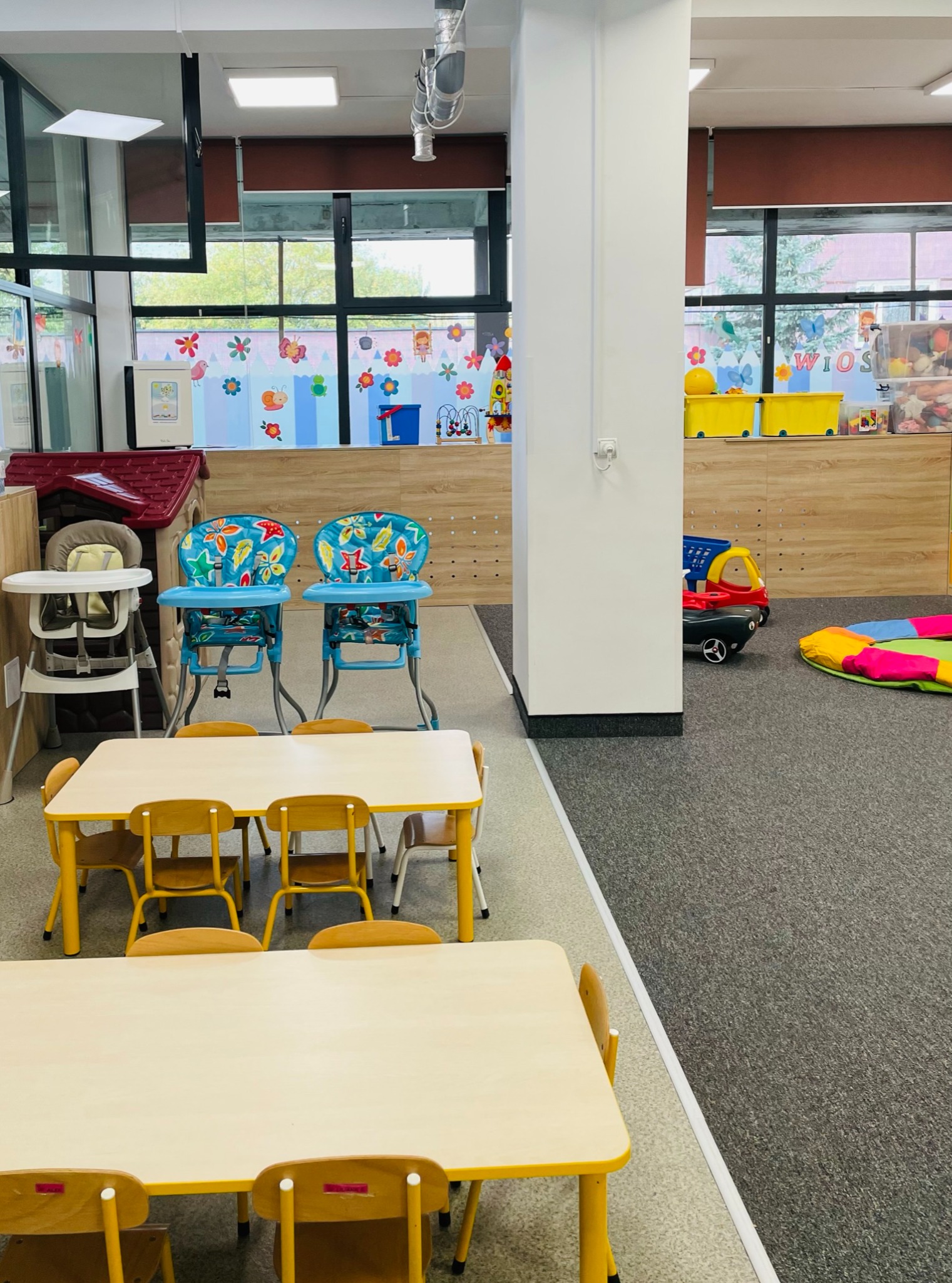 Jasne wnętrze żłobka z żółtymi stolikami i krzesłami dla dzieci, dwoma wysokimi krzesełkami do karmienia oraz dekoracjami ściennymi z motywami kwiatów i zwierząt.