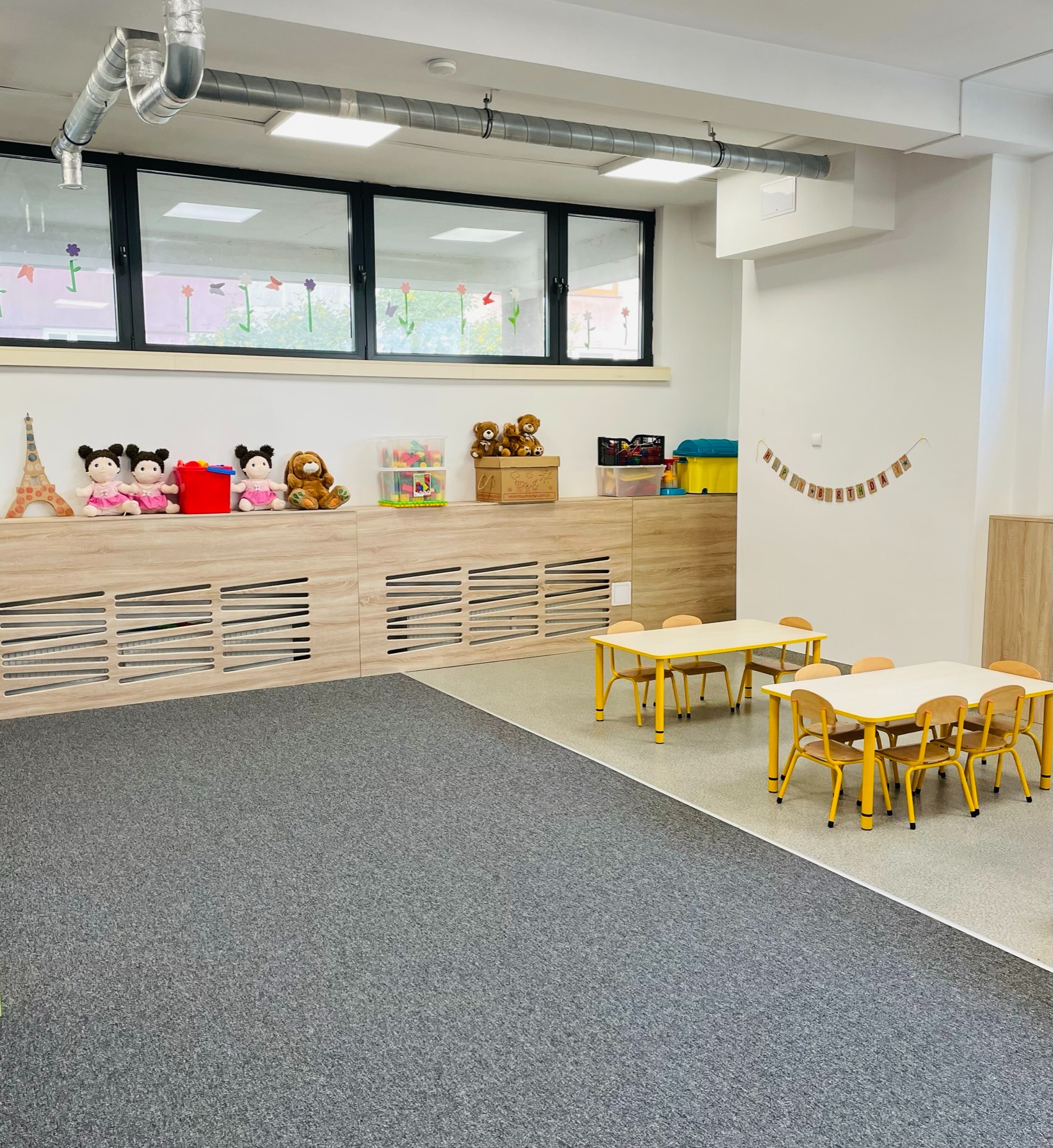 Jasne, przestronne wnętrze sali zabaw z szarym dywanem, niskimi szafkami z zabawkami, stolikami z żółtymi nogami i krzesłami, okno z naklejonymi kwiatkami i rurami wentylacyjnymi pod sufitem.
