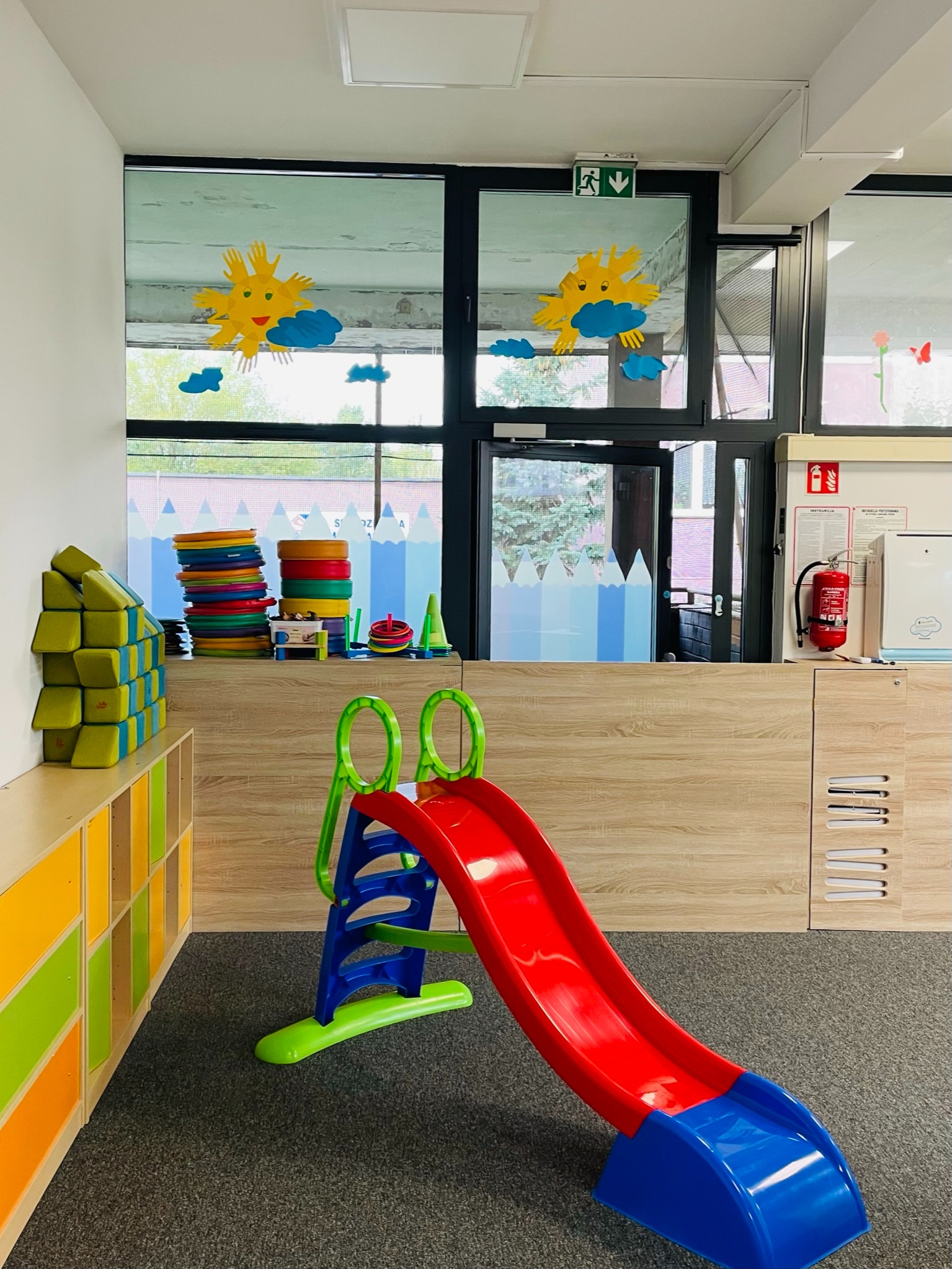 Wnętrze sali zabaw: kolorowa zjeżdżalnia, regał z zabawkami edukacyjnymi, okna ozdobione wycinankami słońca i chmur.